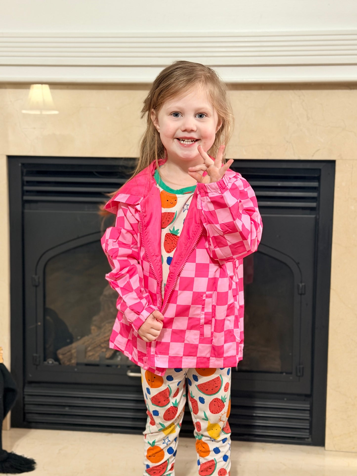 Child wearing a pink checkered jacket and colorful pants in front of a fireplace.