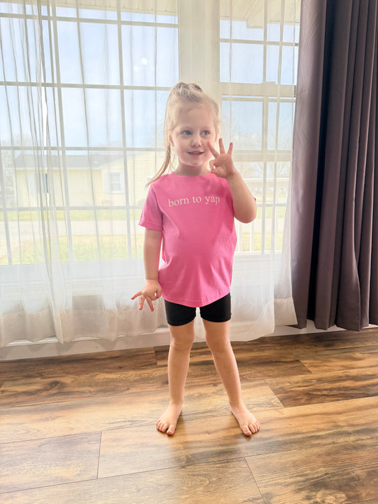 Young girl in a pink shirt standing on a wooden floor with large windows in the background
