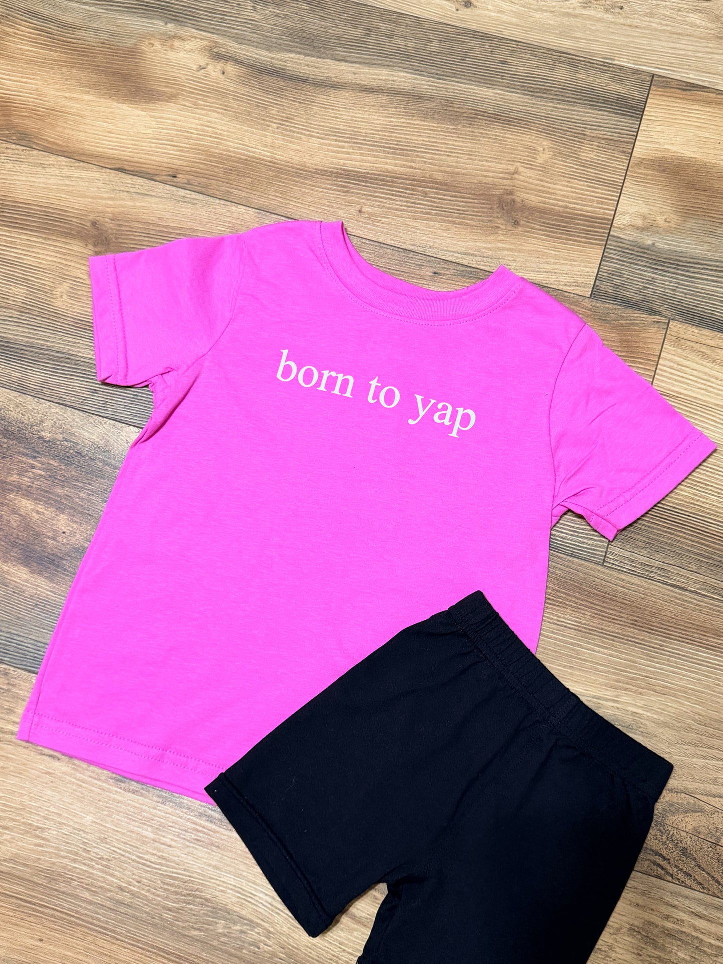 Pink t-shirt with 'born to yap' text and black shorts on a wooden floor.