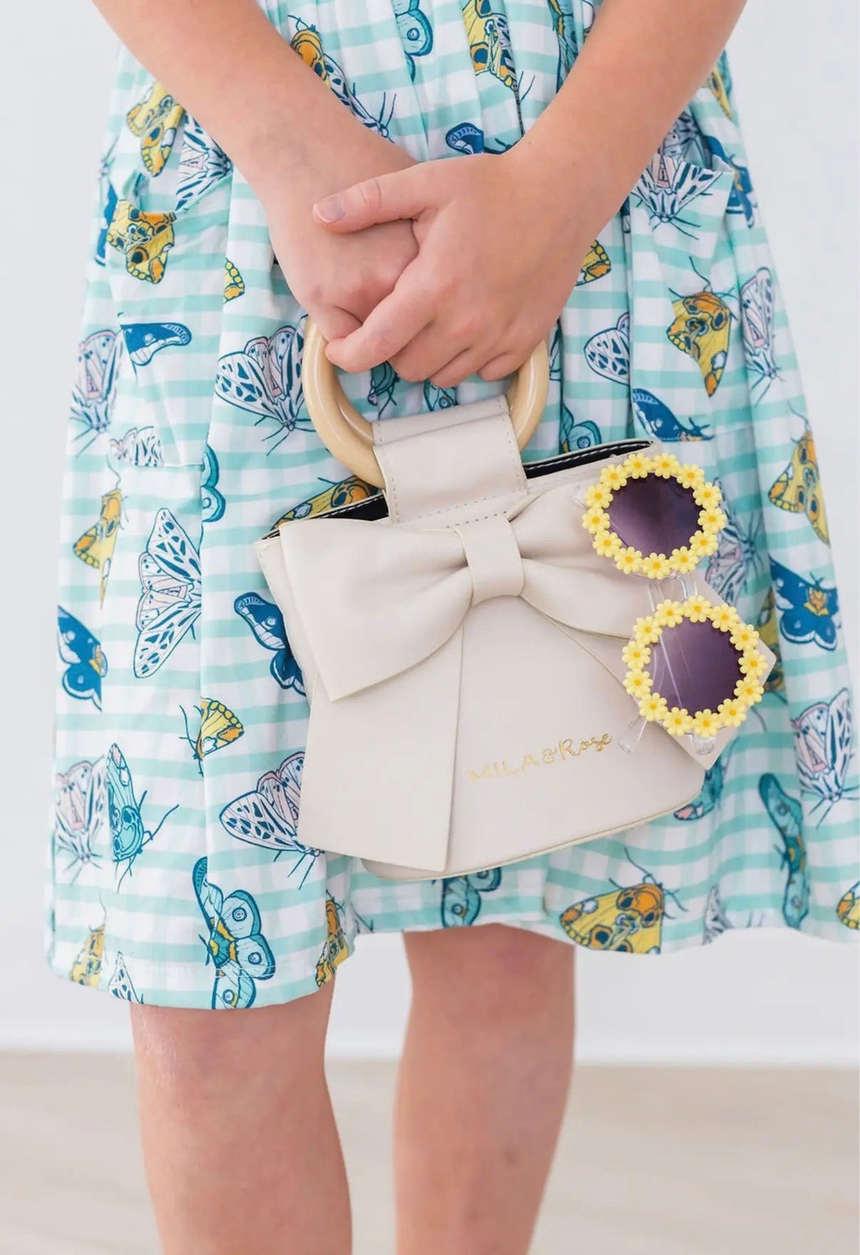 Person wearing a blue dress with butterfly pattern holding a beige handbag with bow detail and yellow daisy sunglasses.