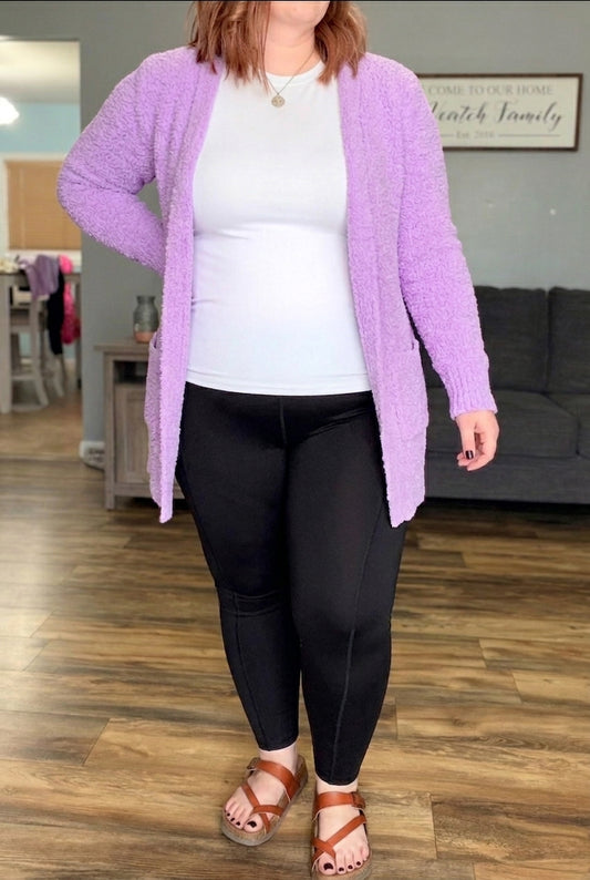Person wearing a purple cardigan, white shirt, black pants, and brown sandals in a living room.