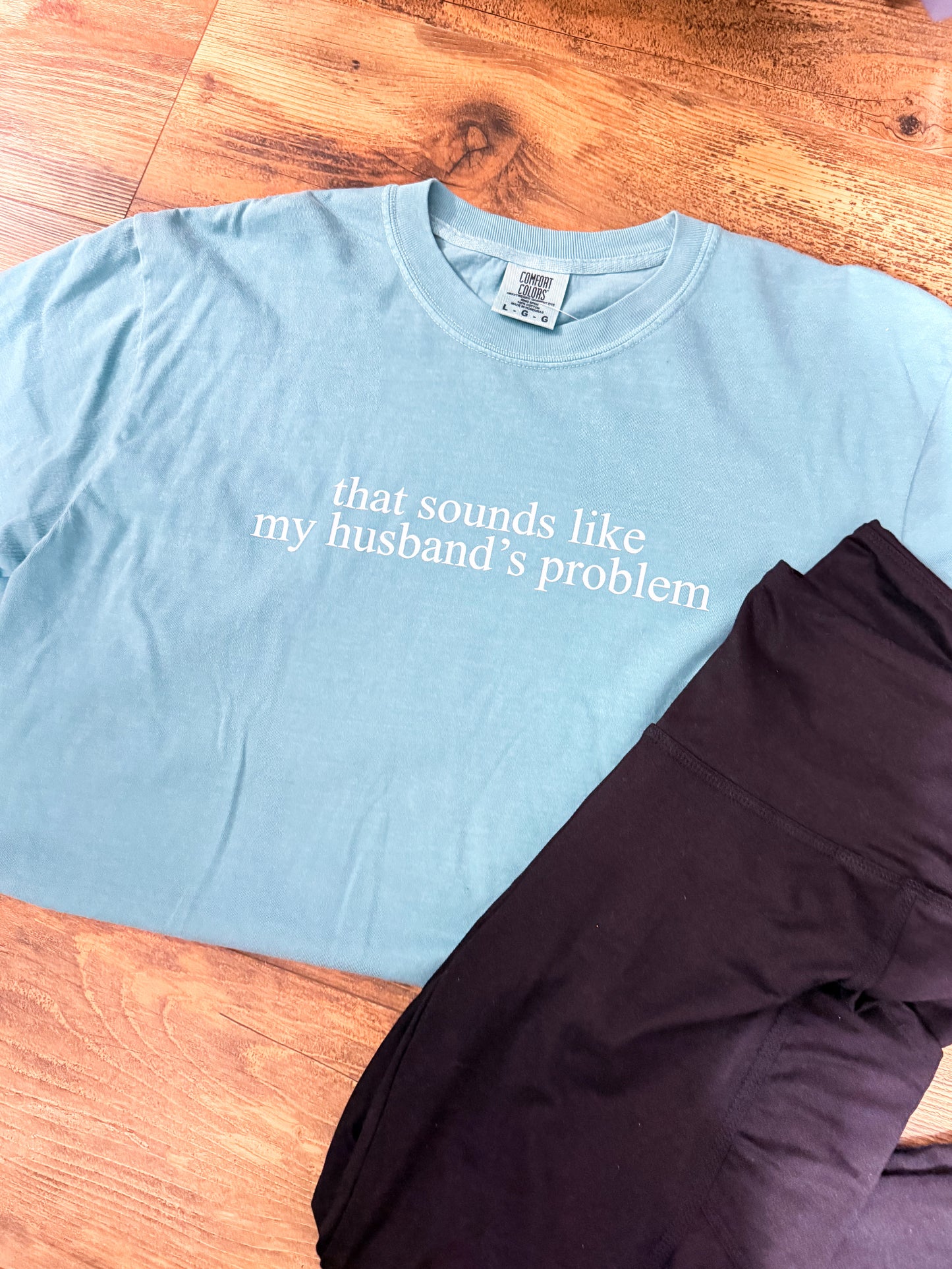 Light blue t-shirt with text 'that sounds like my husband's problem' on a wooden surface