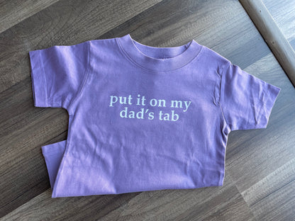 Purple t-shirt with text 'put it on my dad's tab' on a wooden surface