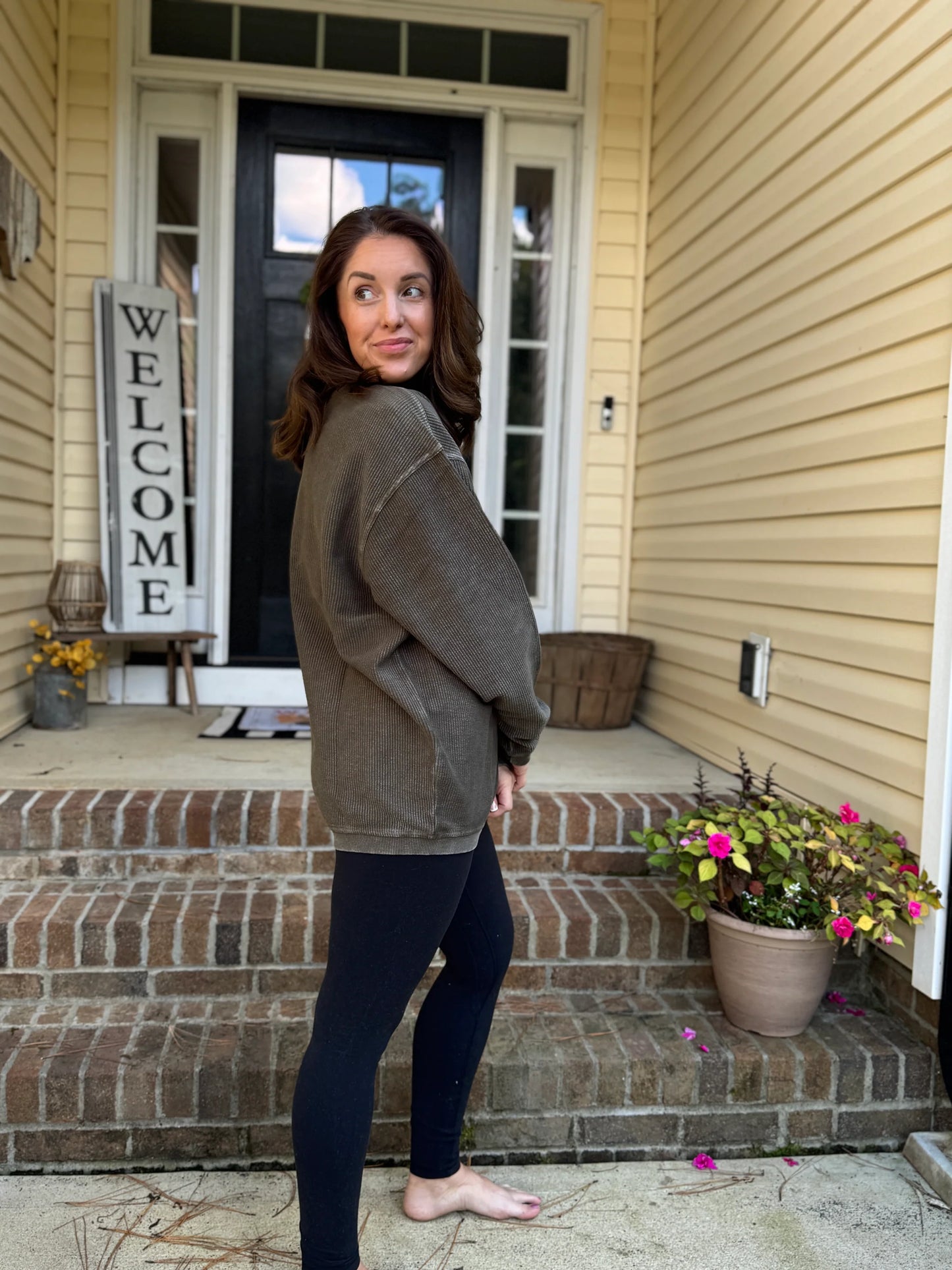 Woman standing on a porch with a 'Welcome' sign and potted plants. Woman is wearing a hunter green corded crew and black leggings