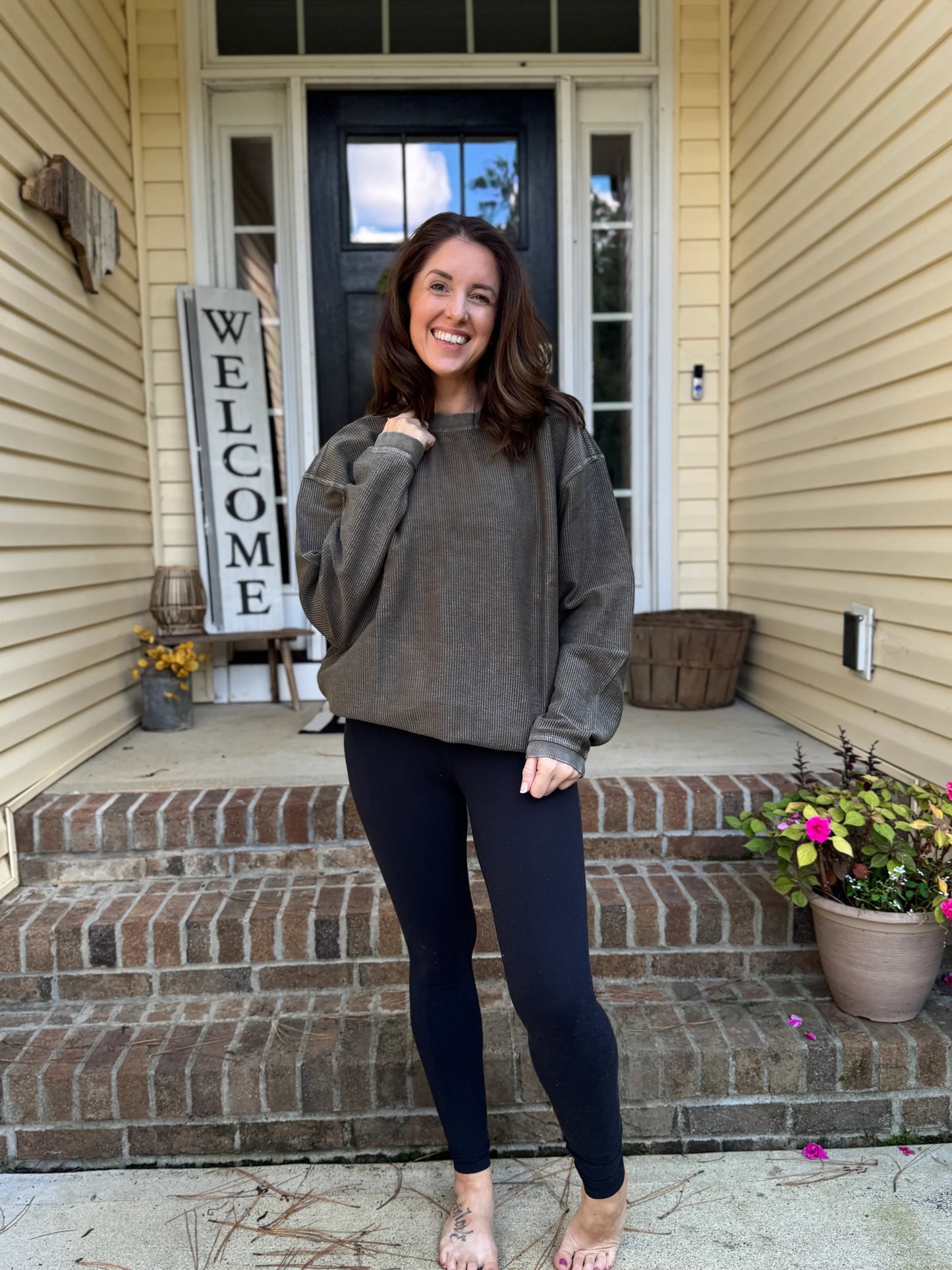Woman standing on a porch with a 'Welcome' sign and potted plants. Woman is wearing a hunter green corded crew and black leggings
