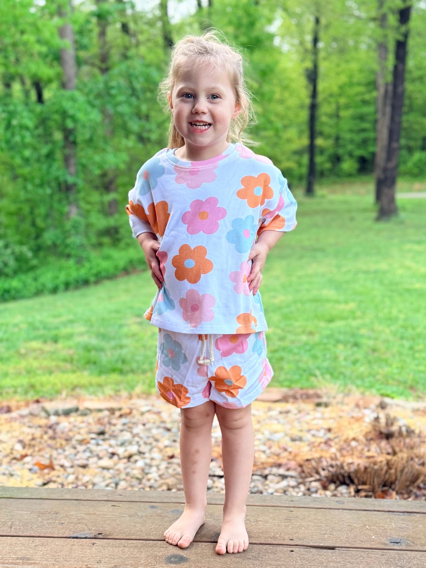 Young girl wearing a floral outfit standing on a wooden deck with greenery in the background