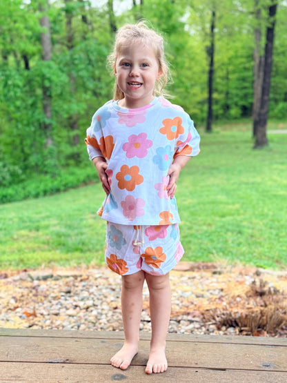 Young girl wearing a floral outfit standing on a wooden deck with greenery in the background