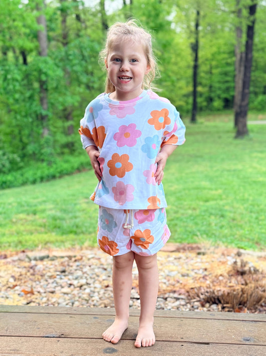 Young girl wearing a floral outfit standing on a wooden deck with greenery in the background