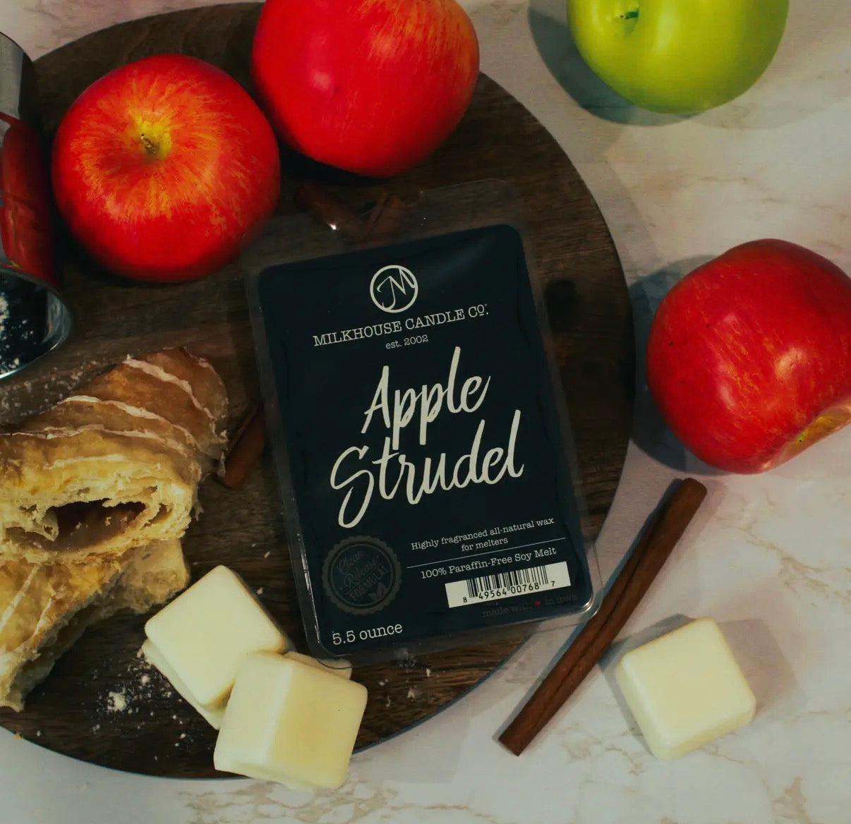Milkhouse Candle Co. Apple Strudel candle on a wooden board with apples, butter, and cinnamon sticks.