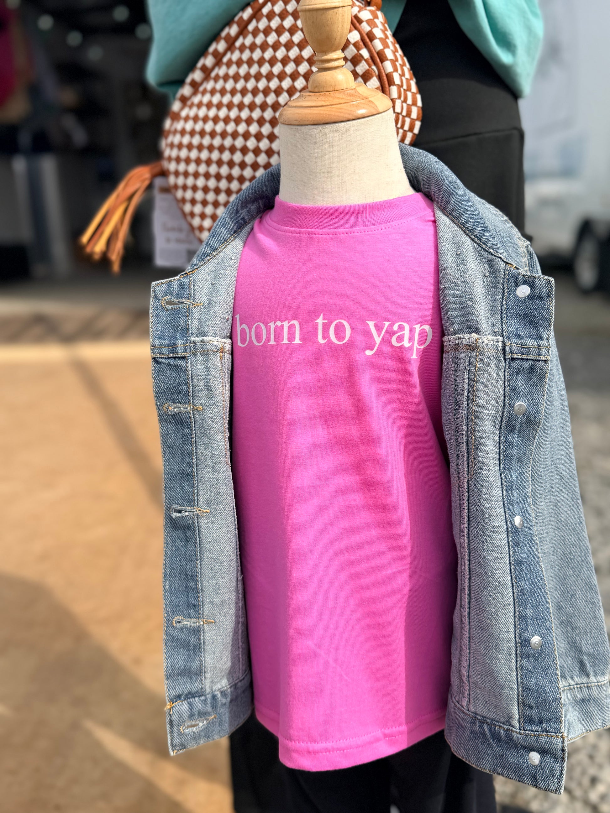 Mannequin wearing a pink 'Born to yap' shirt with a denim jacket outdoors.