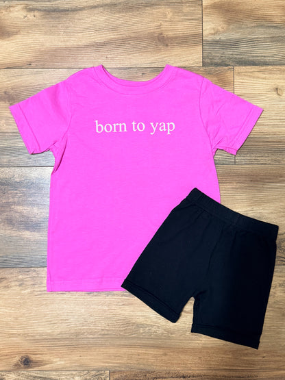 Pink t-shirt with 'born to yap' text and black shorts on a wooden surface