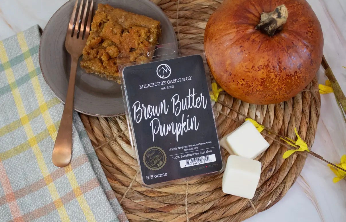Candle labeled 'Brown Butter Pumpkin' from Milkhouse Candle Co. on a woven mat with a slice of cake and a small pumpkin.