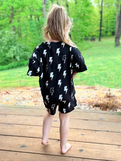 Child wearing a black outfit with lightning bolt patterns standing on a wooden deck with a green outdoor background.