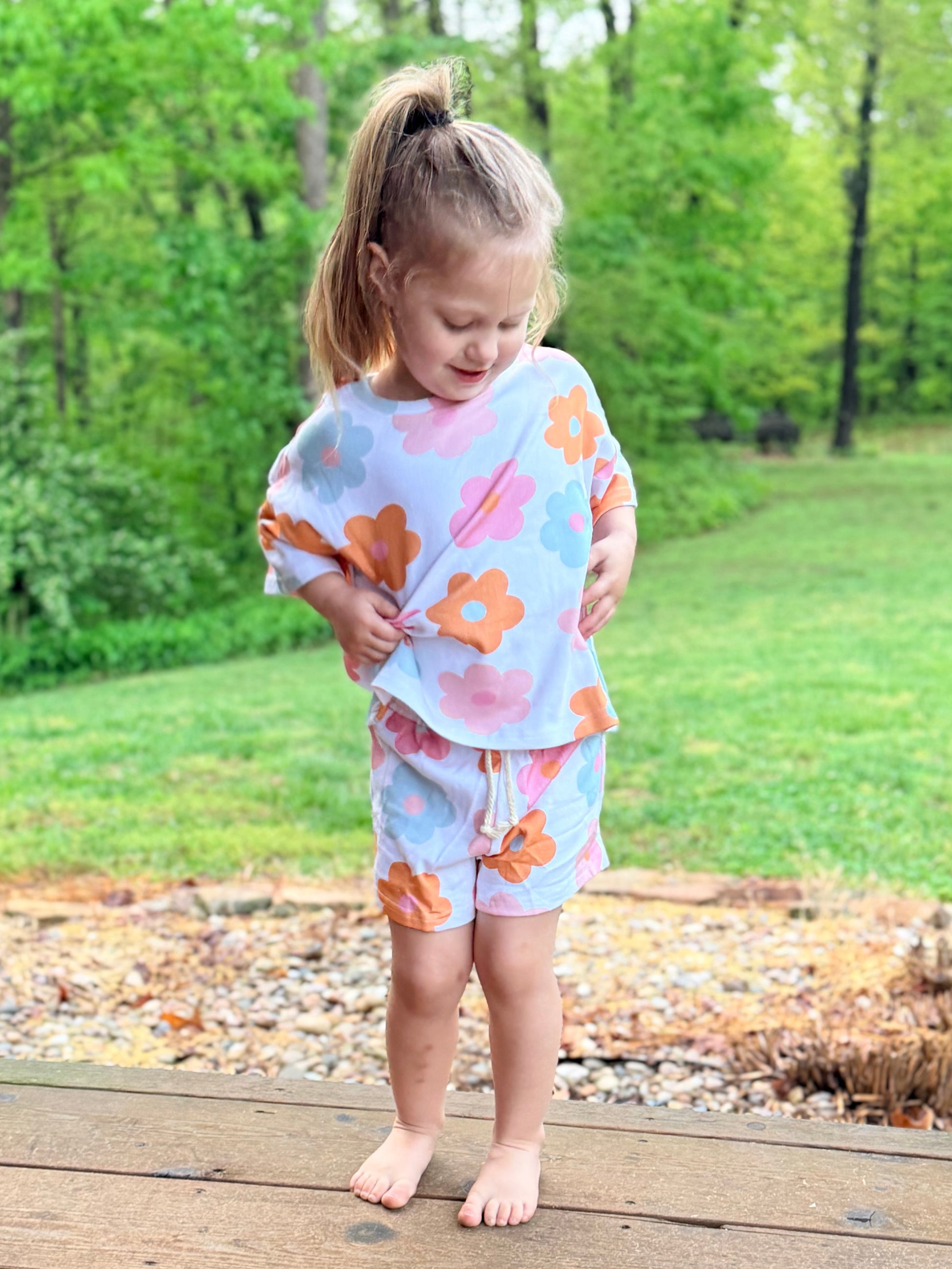 Child in a floral outfit standing on a wooden deck with a green, leafy background