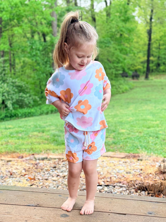 Child in a floral outfit standing on a wooden deck with a green, leafy background