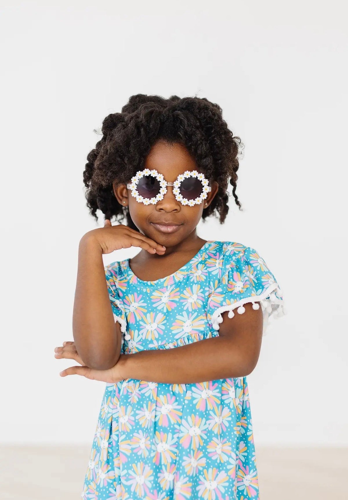Young girl wearing a blue floral dress and sunglasses with white daisies on a light gray background