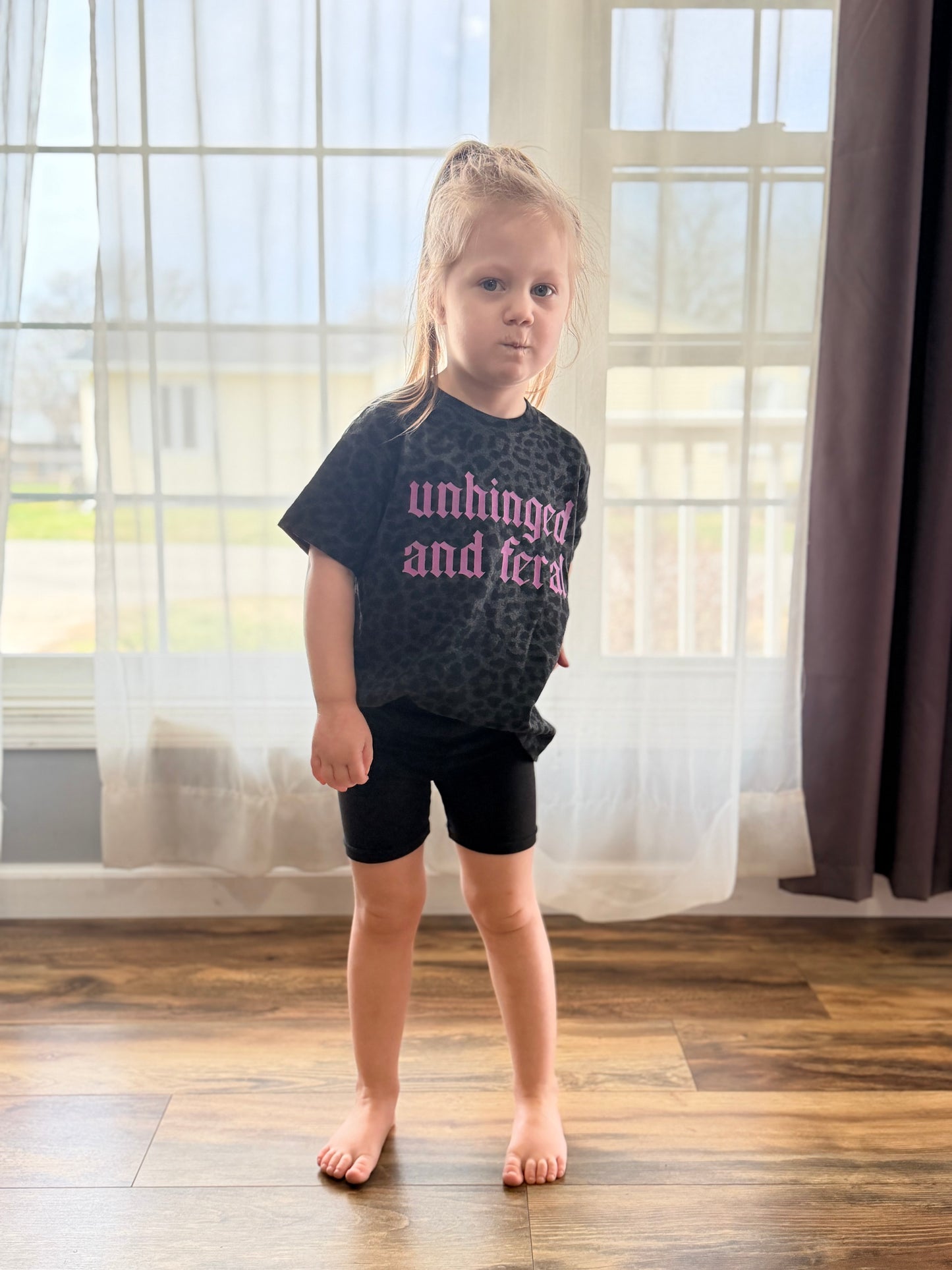 Young girl wearing a black t-shirt with text, standing in a room with large windows.