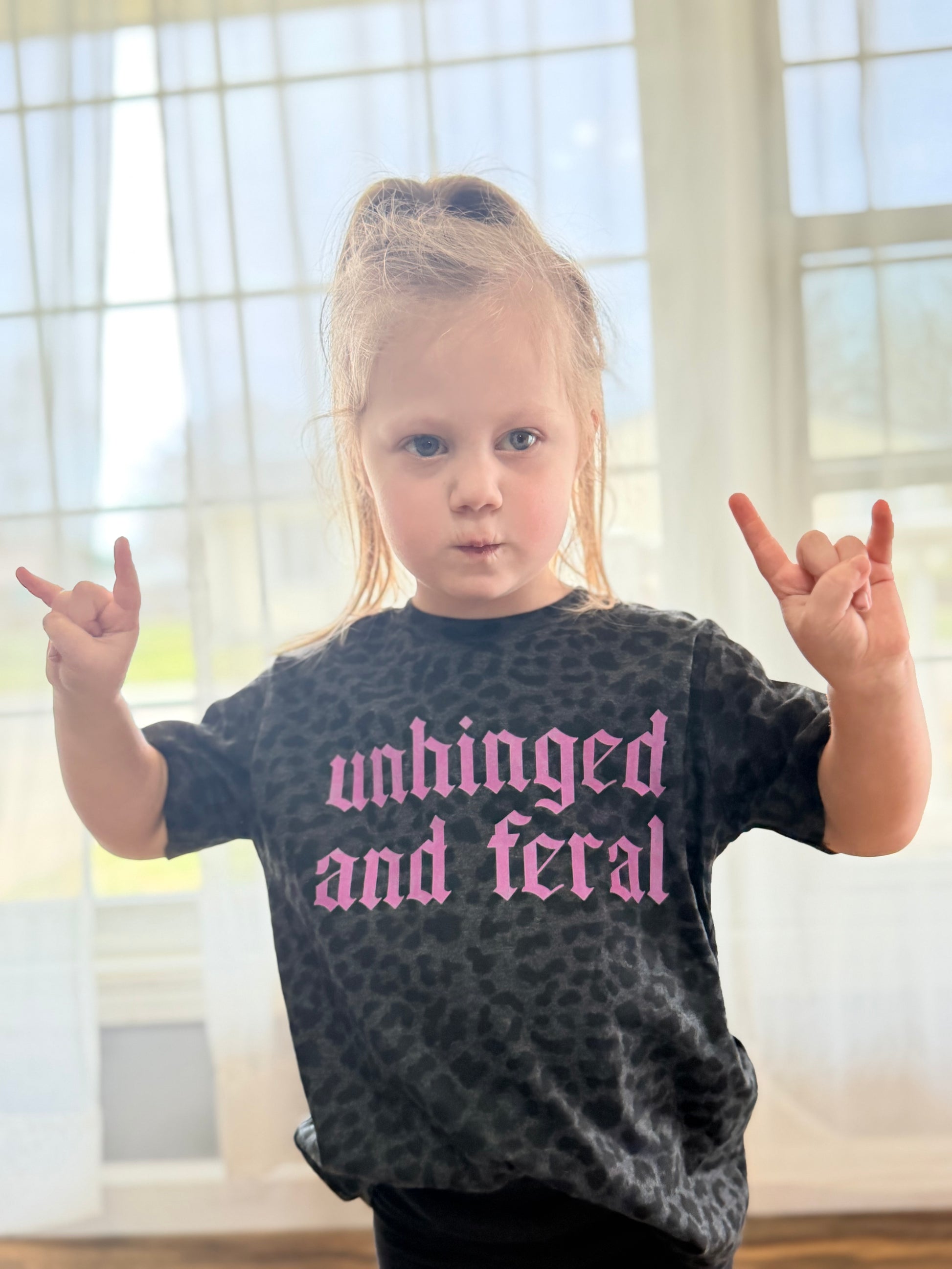 Child wearing a shirt with 'unhinged and feral' text, making hand gestures indoors.
