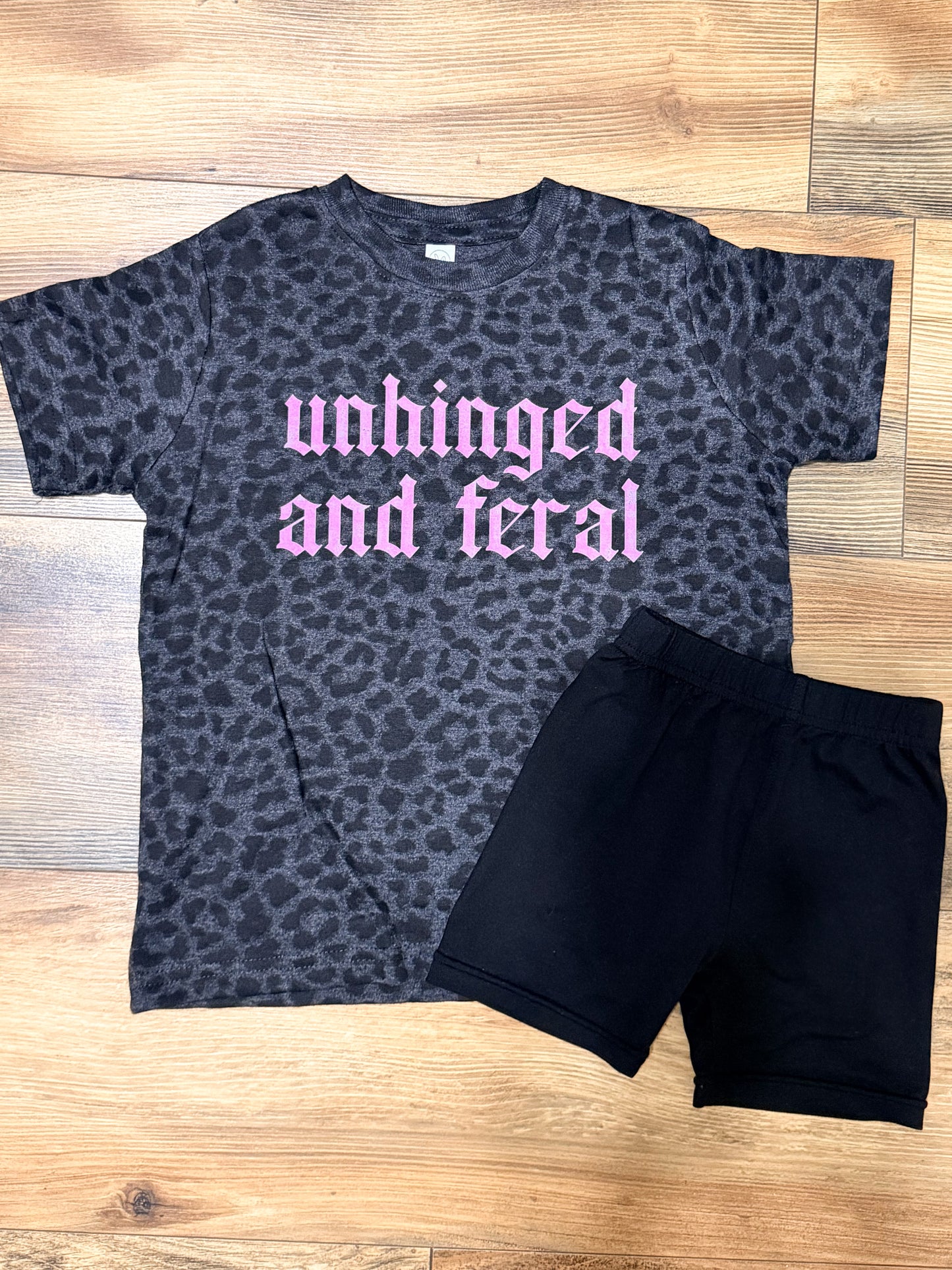 Gray leopard print t-shirt with pink text and black shorts on a wooden floor.