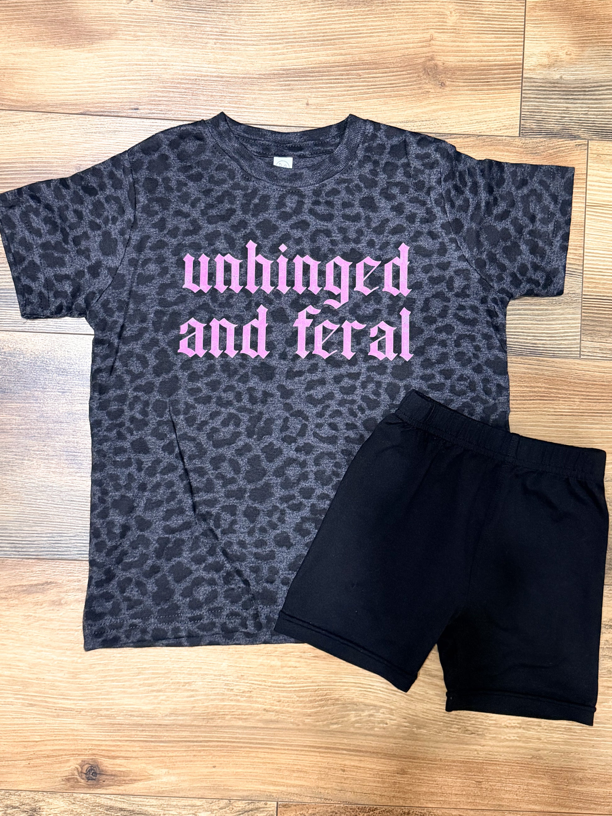 Gray leopard print t-shirt with pink text and black shorts on a wooden floor.