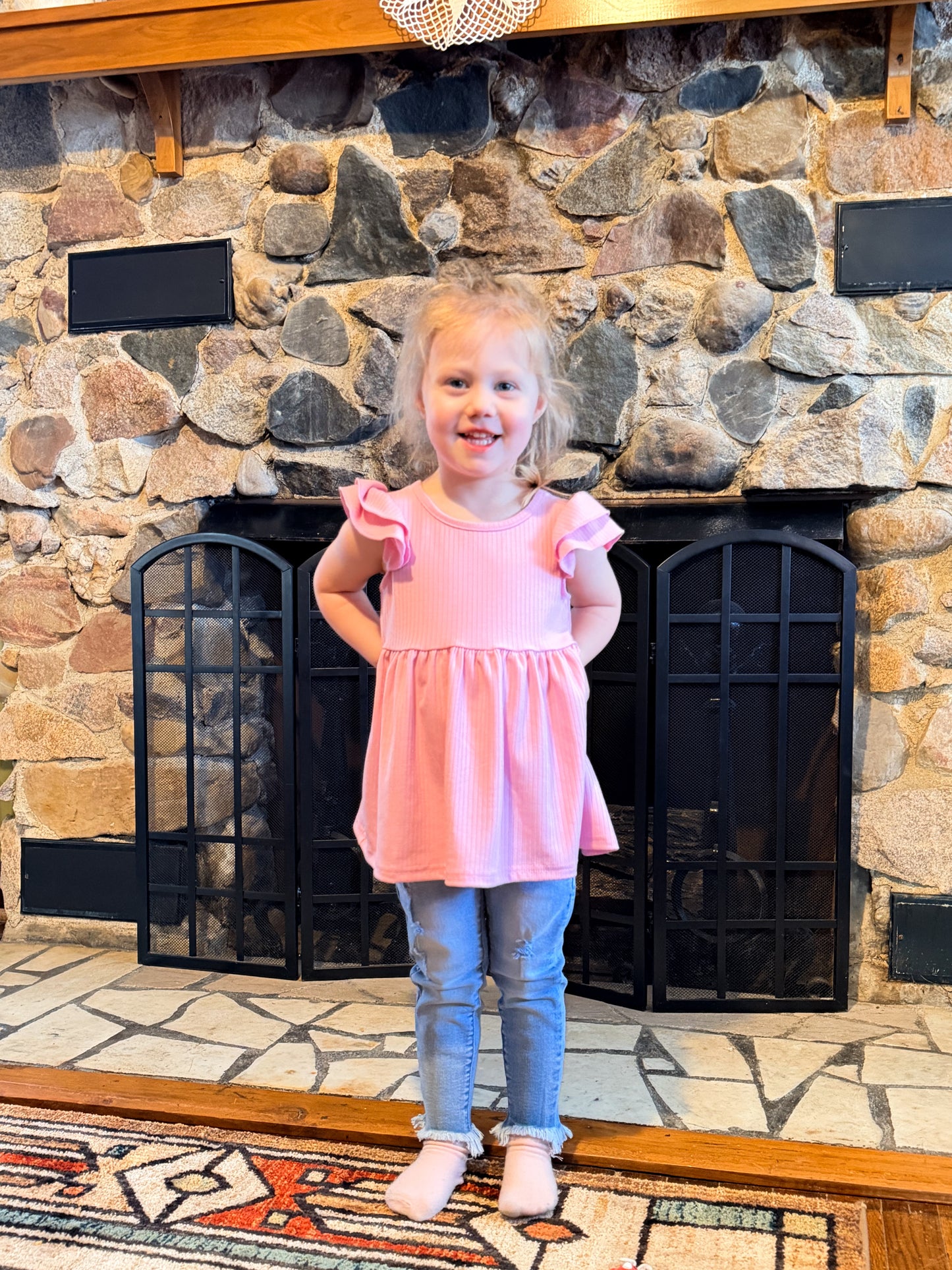 Young girl in a pink shirt standing in front of a stone fireplace.