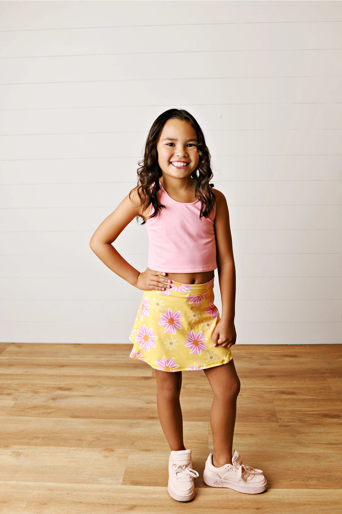 Young girl wearing a pink top and yellow floral skirt on a wooden floor.
