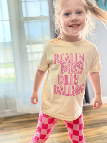 Child wearing a cream t-shirt with pink text and pink checkered pants indoors.