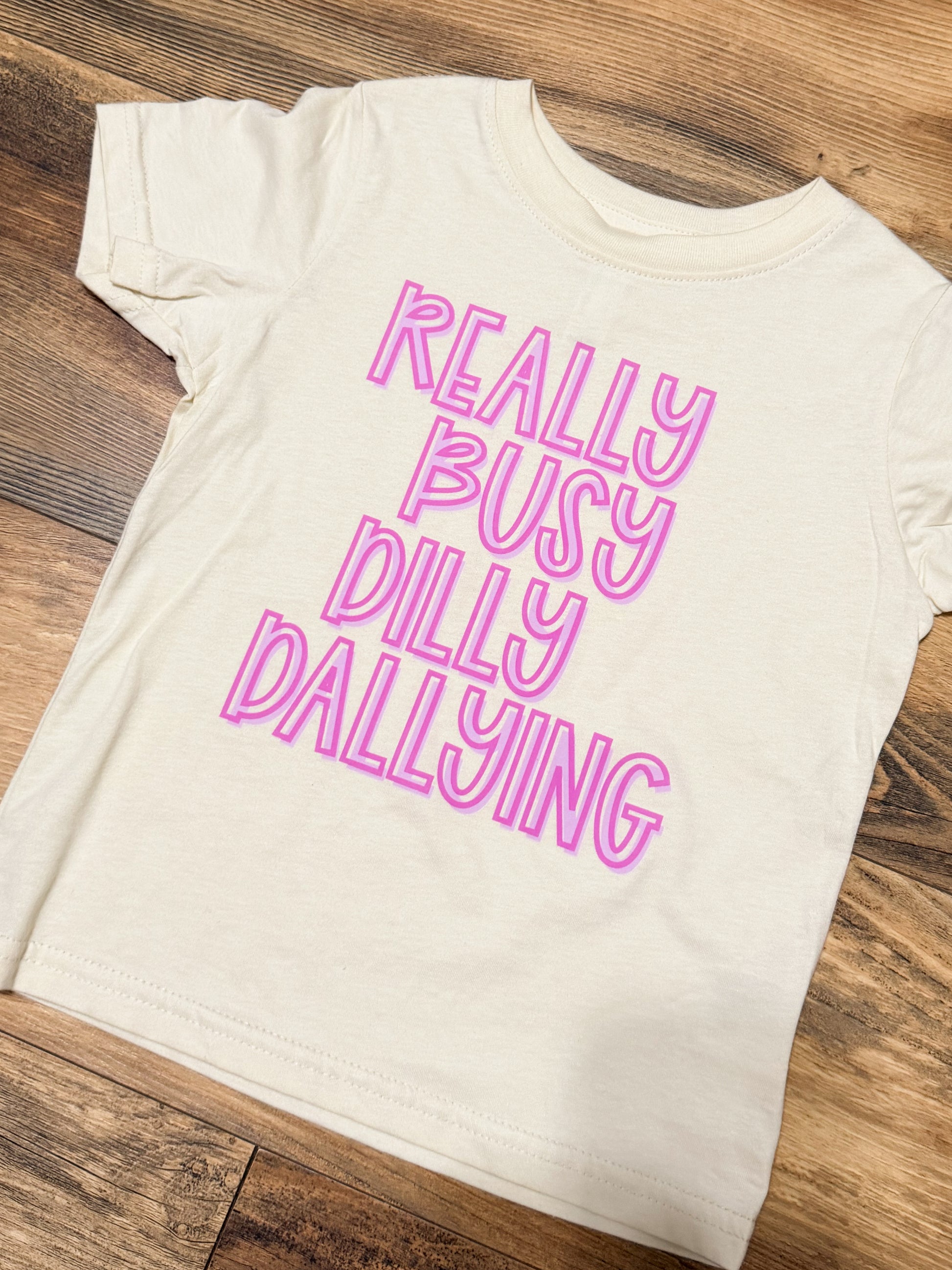 White t-shirt with pink text 'REALLY BUSY DILLY DALLIYING' on a wooden surface