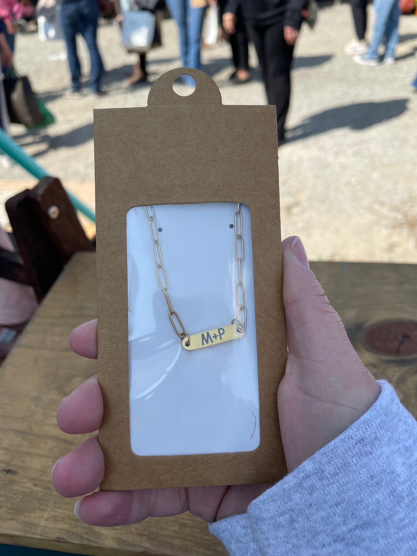 Gold necklace with 'M+P' engraving in a brown packaging holder, held by a person outdoors.