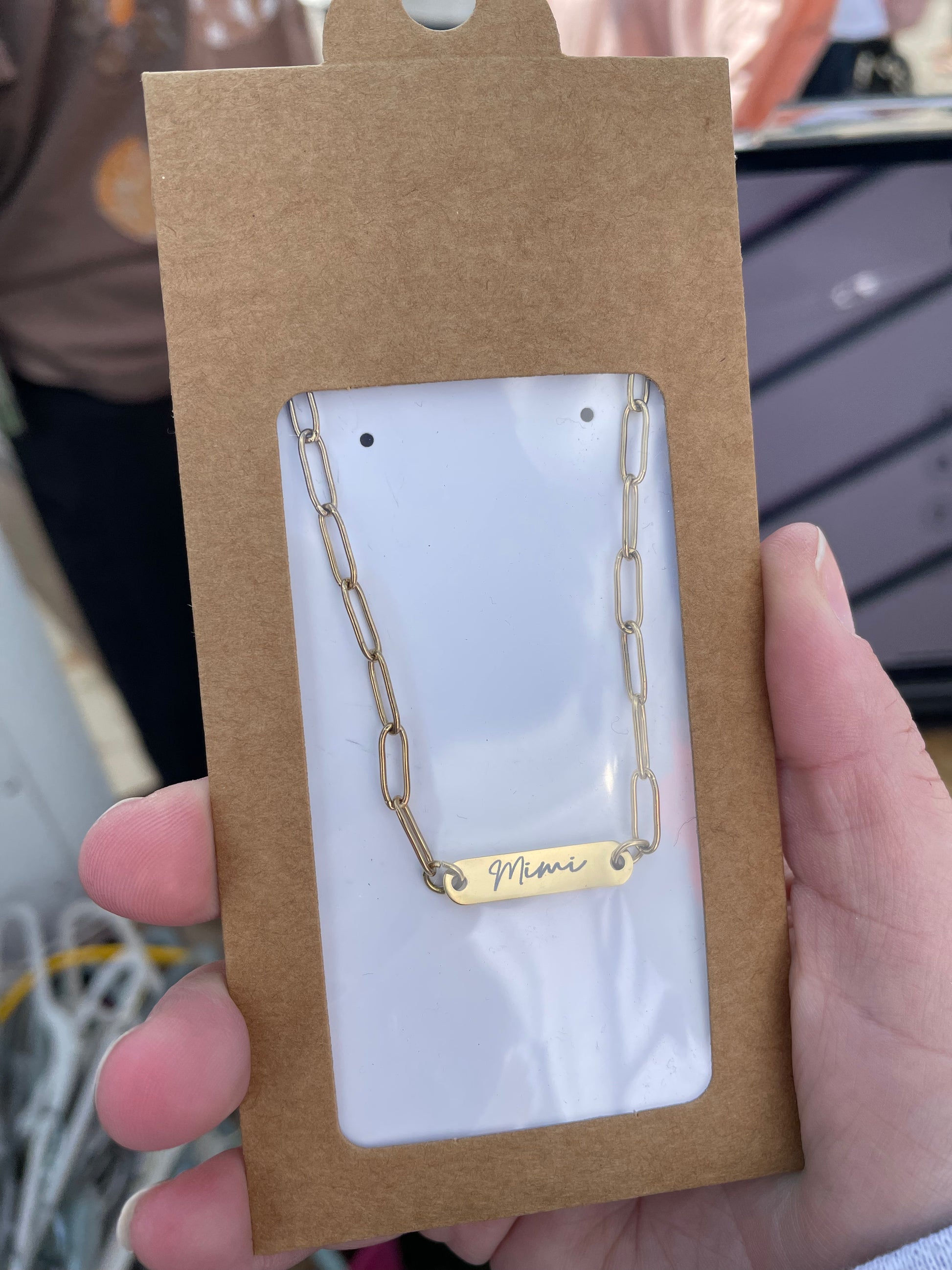 Gold necklace with 'Mimi' engraving on a brown card held by a hand.