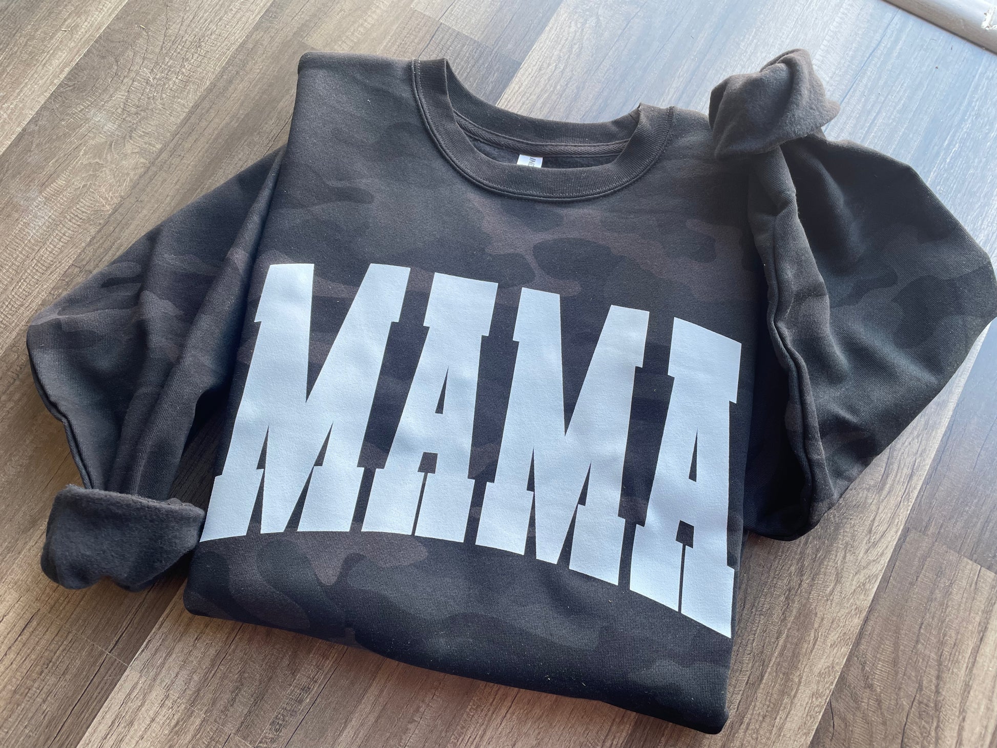 Black camo sweatshirt with 'MAMA' printed on it laid flat on a wooden surface