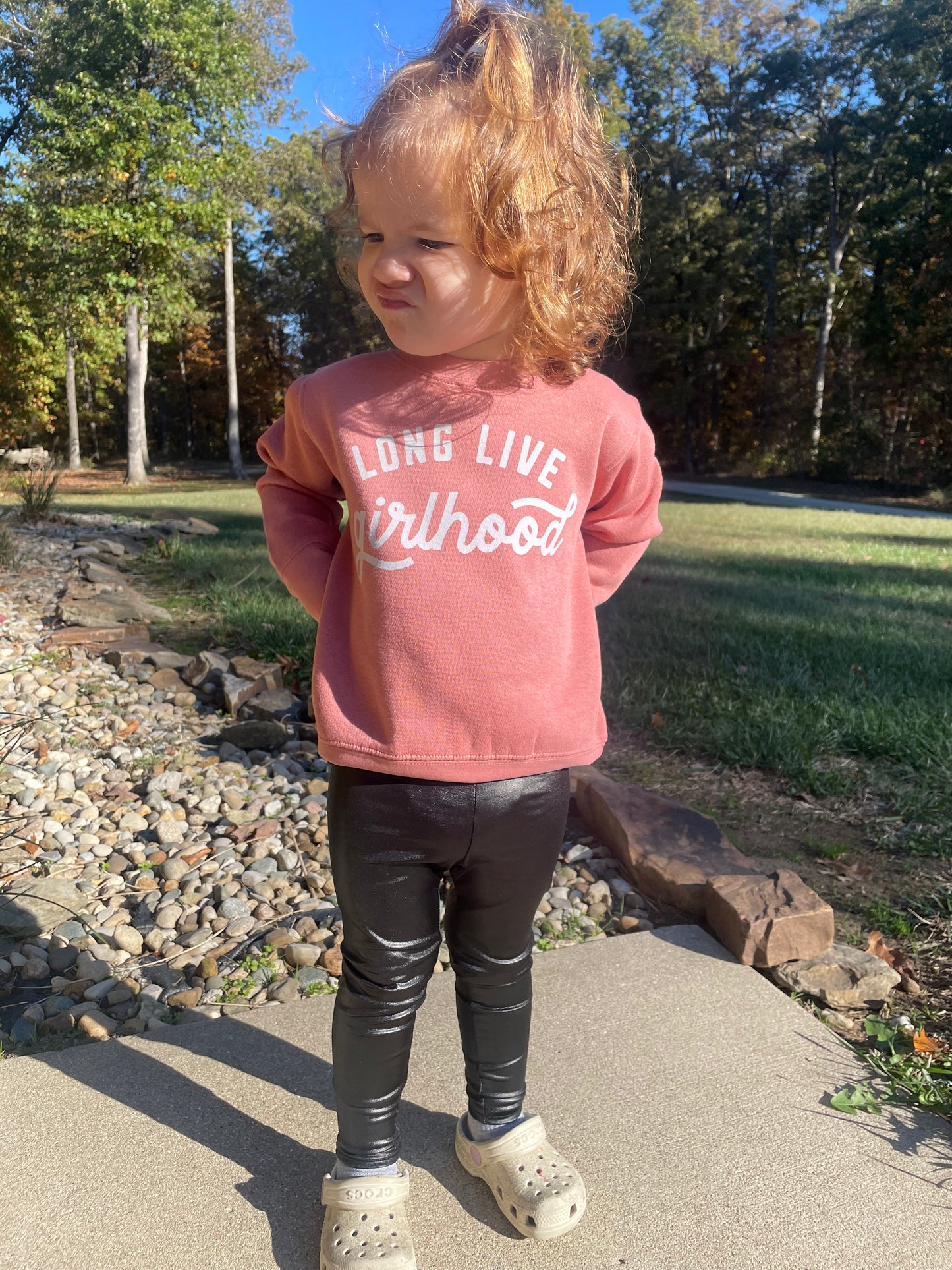 Child wearing a pink sweatshirt with text, standing outdoors on a sunny day.