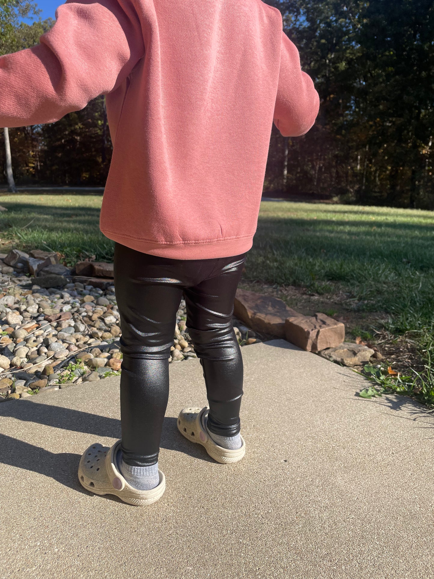 Person wearing a pink sweatshirt and black leggings standing on a sidewalk with grass and trees in the background.