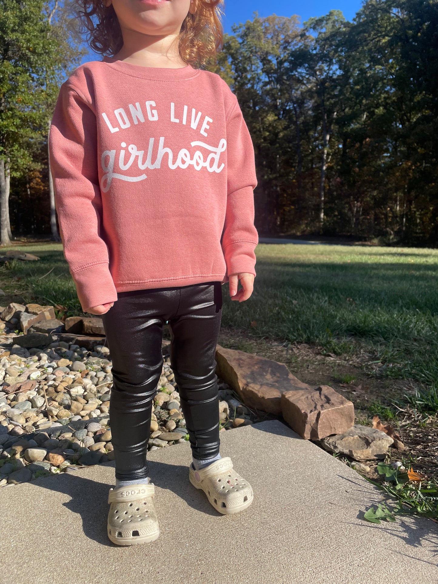 Child wearing a pink sweatshirt with text, black pants, and white Crocs standing outdoors.