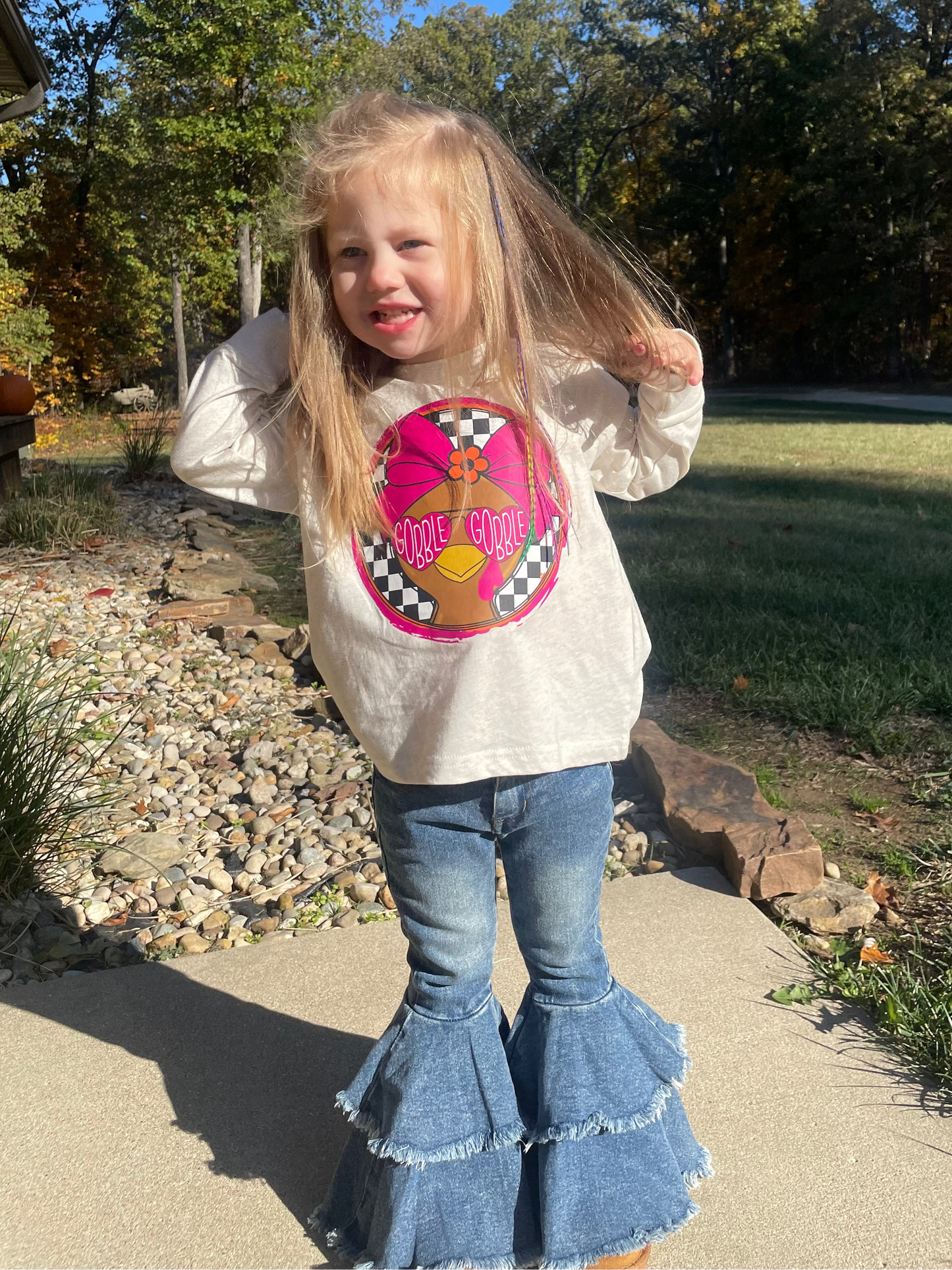 Child wearing a thanksgiving graphic long sleeve and blue jeans outdoors.