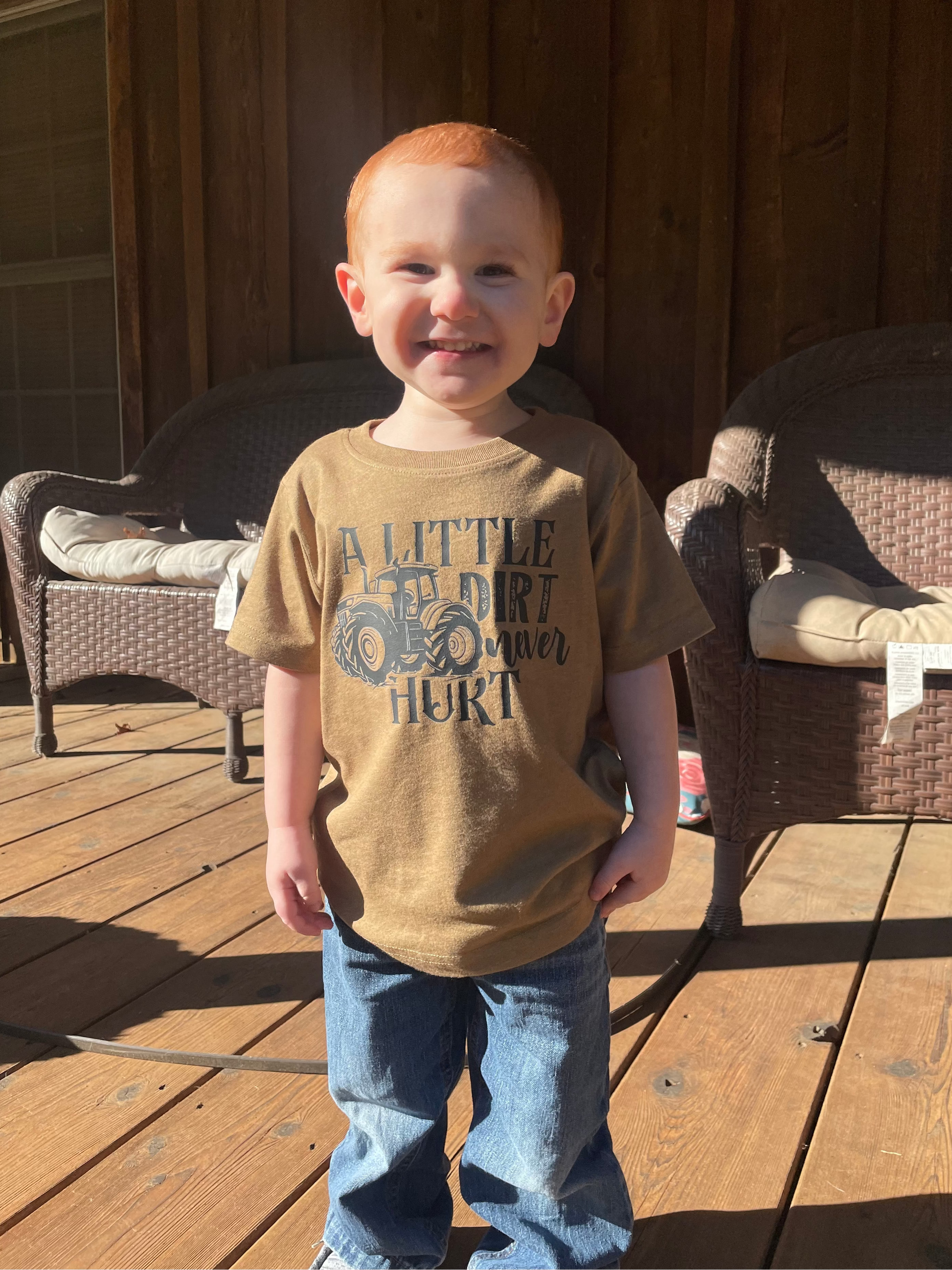 Child wearing a brown shirt with text standing on a wooden deck.
