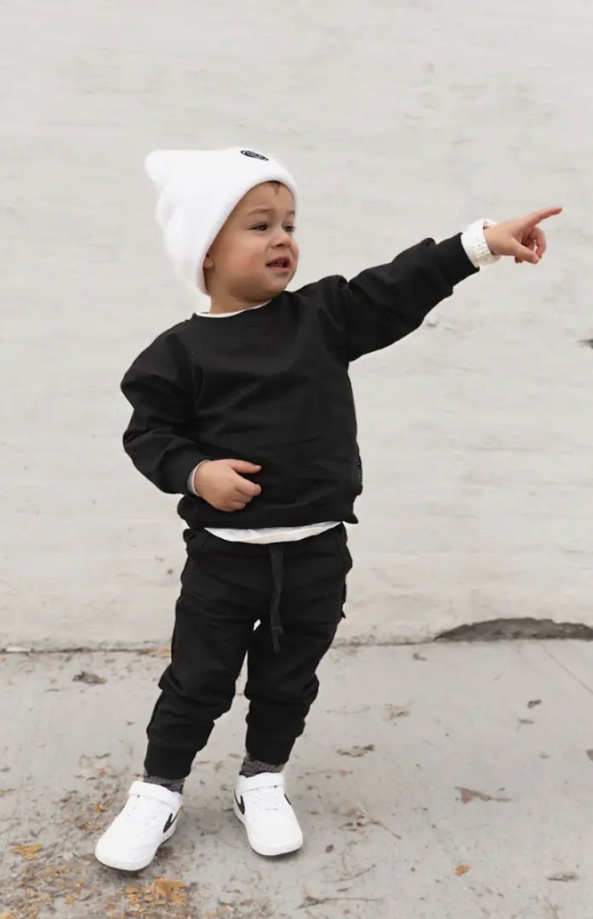 Child wearing a black outfit with white shoes and a white beanie, pointing towards something.