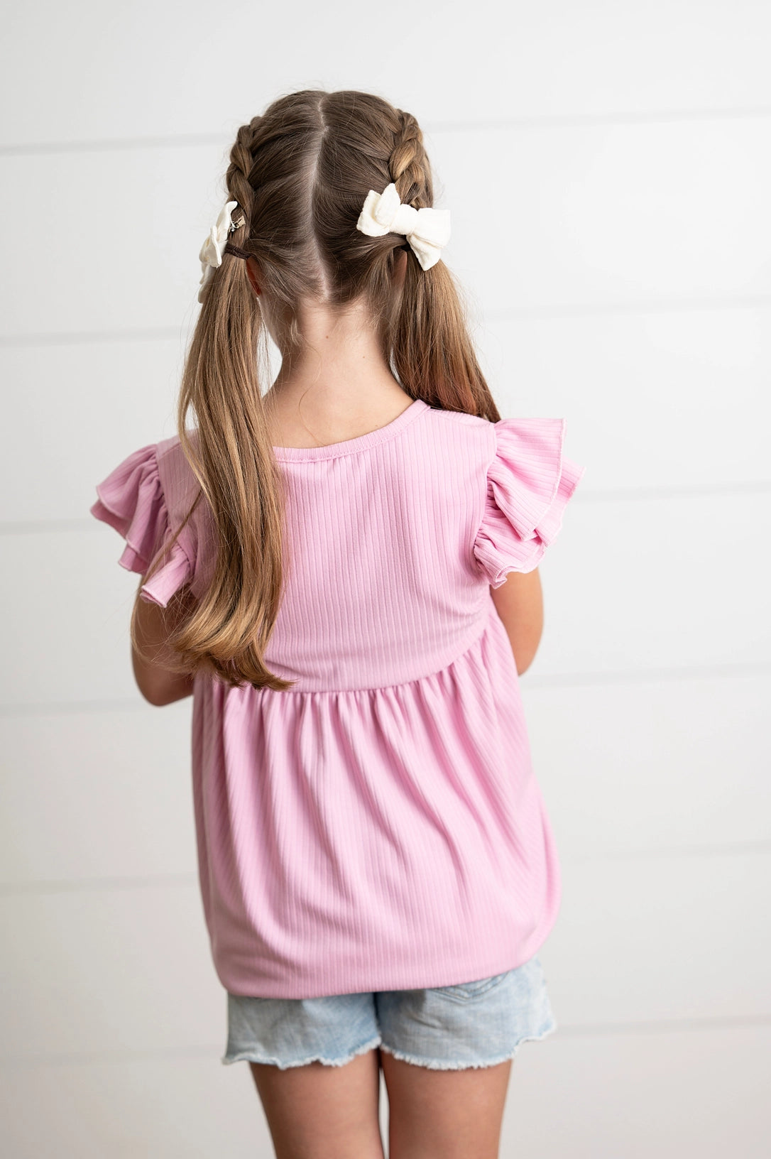 Young girl wearing a pink shirt with ruffle sleeves and denim shorts, standing against a light background.