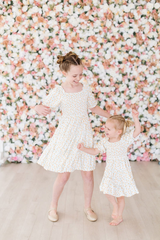Two girls in matching white dresses with a marigold print standing in front of a floral wall.
