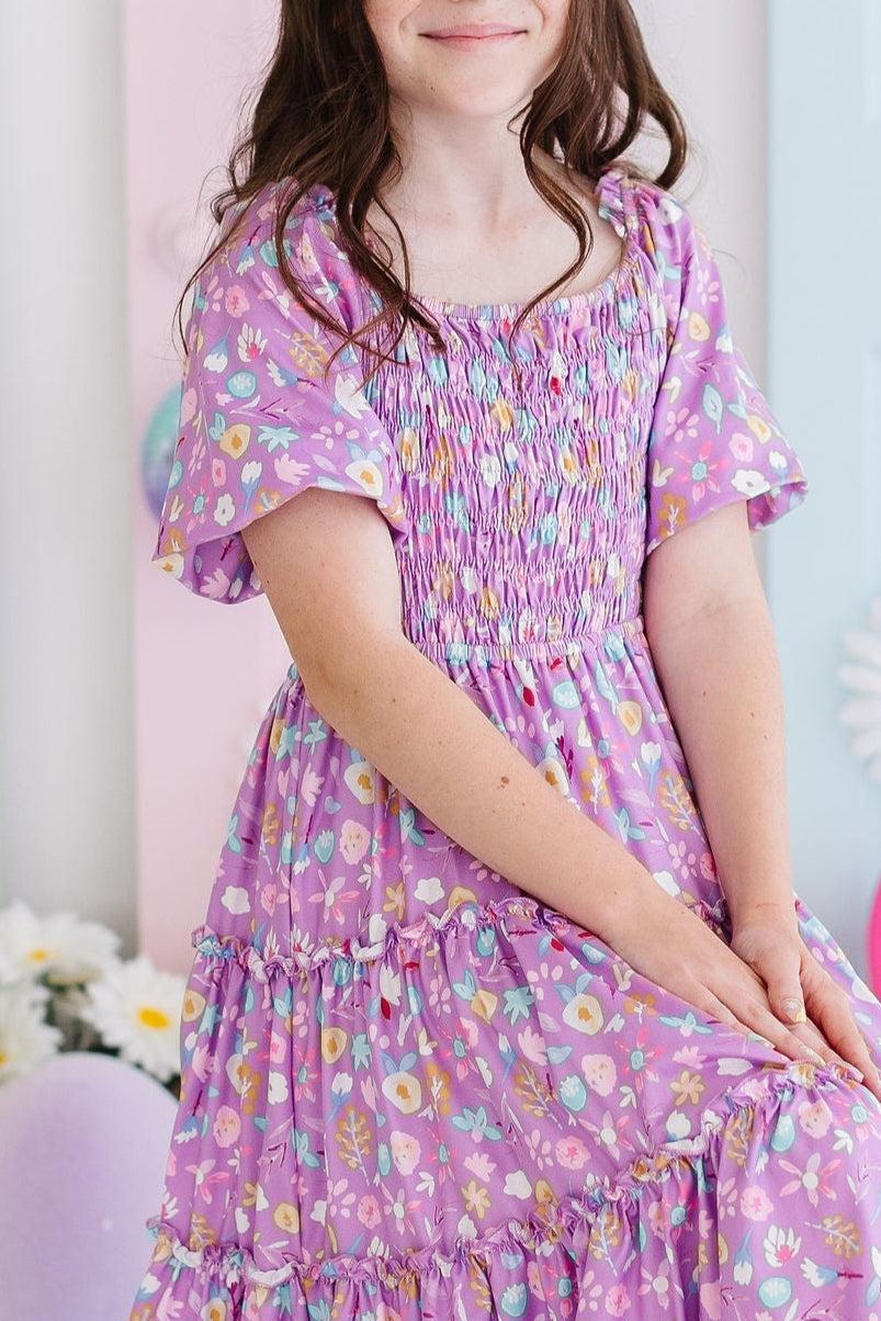 Child wearing a purple floral dress sitting against a pink background