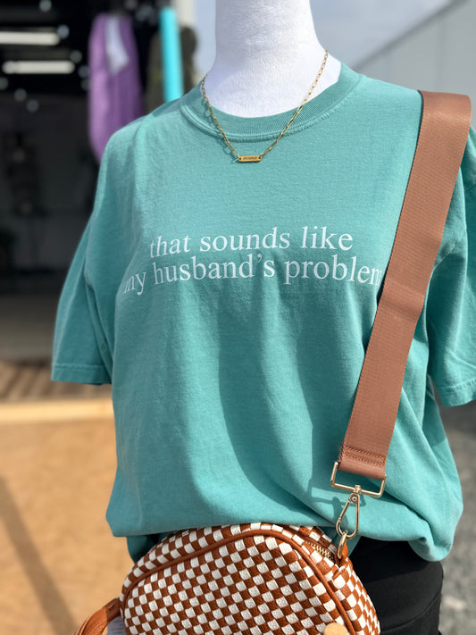 Mannequin wearing a teal t-shirt with text saying "that sounds like my husband's problem" and carrying a brown and white checkered bag.