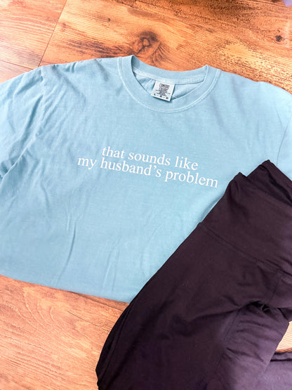 Light blue t-shirt with text 'that sounds like my husband's problem' on a wooden surface