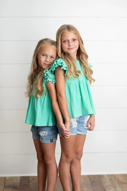 Two young girls wearing matching turquoise tops and denim shorts standing against a white wooden panel background.