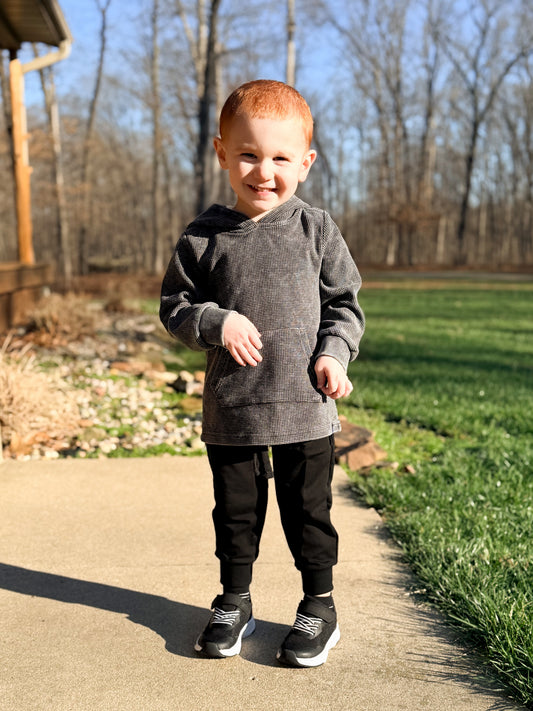 Child wearing a gray sweatshirt and black pants standing on a path with trees in the background